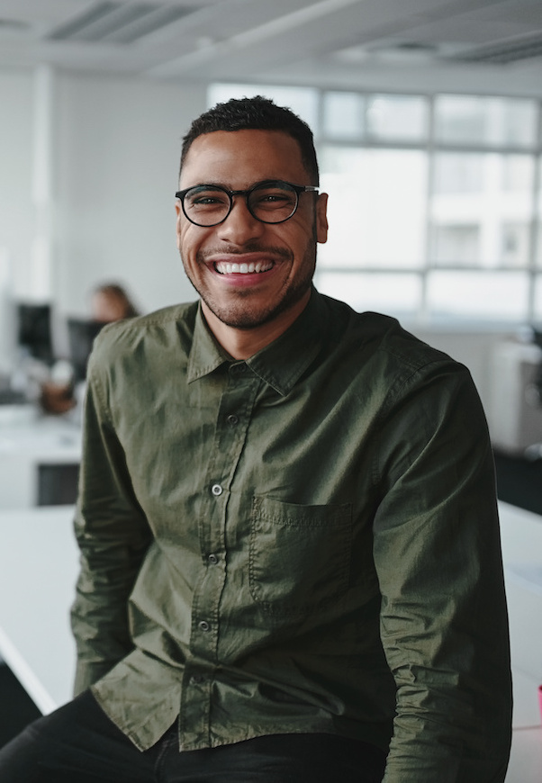 Portrait of a successful smiling young professional businessman sitting over desk in front of colleague working in background